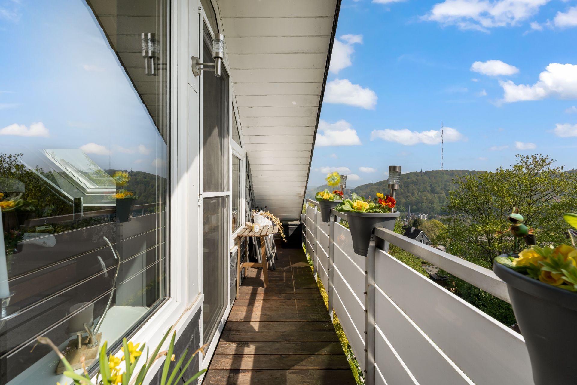 Schmaler Balkon mit Holzboden, Blumenkästen am Geländer und Blick auf grüne Hügel.