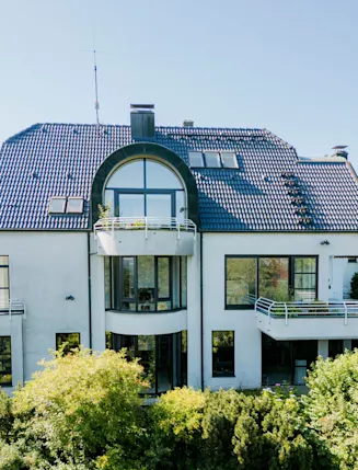 Modernes, weißes Wohnhaus mit dunklem Dach, Balkonen und großen Fenstern, von Grün umgeben.