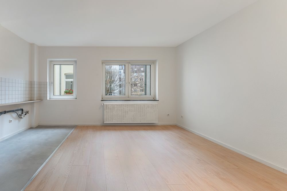 Heller, leerer Raum mit Holzfußboden, zwei Fenstern und Anschlüssen für eine Küche.