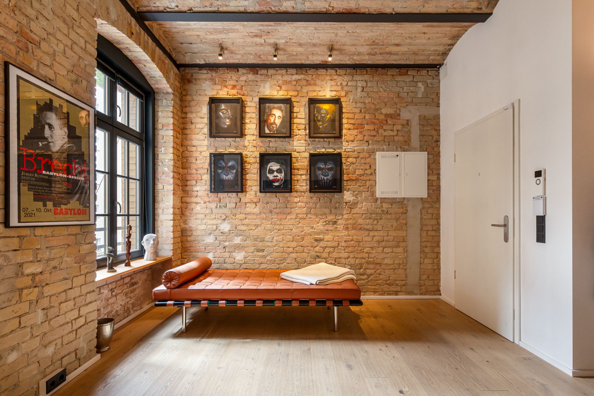 Zimmer mit Backsteinwand, Liege am Fenster, sechs Porträtbildern und Holzboden.