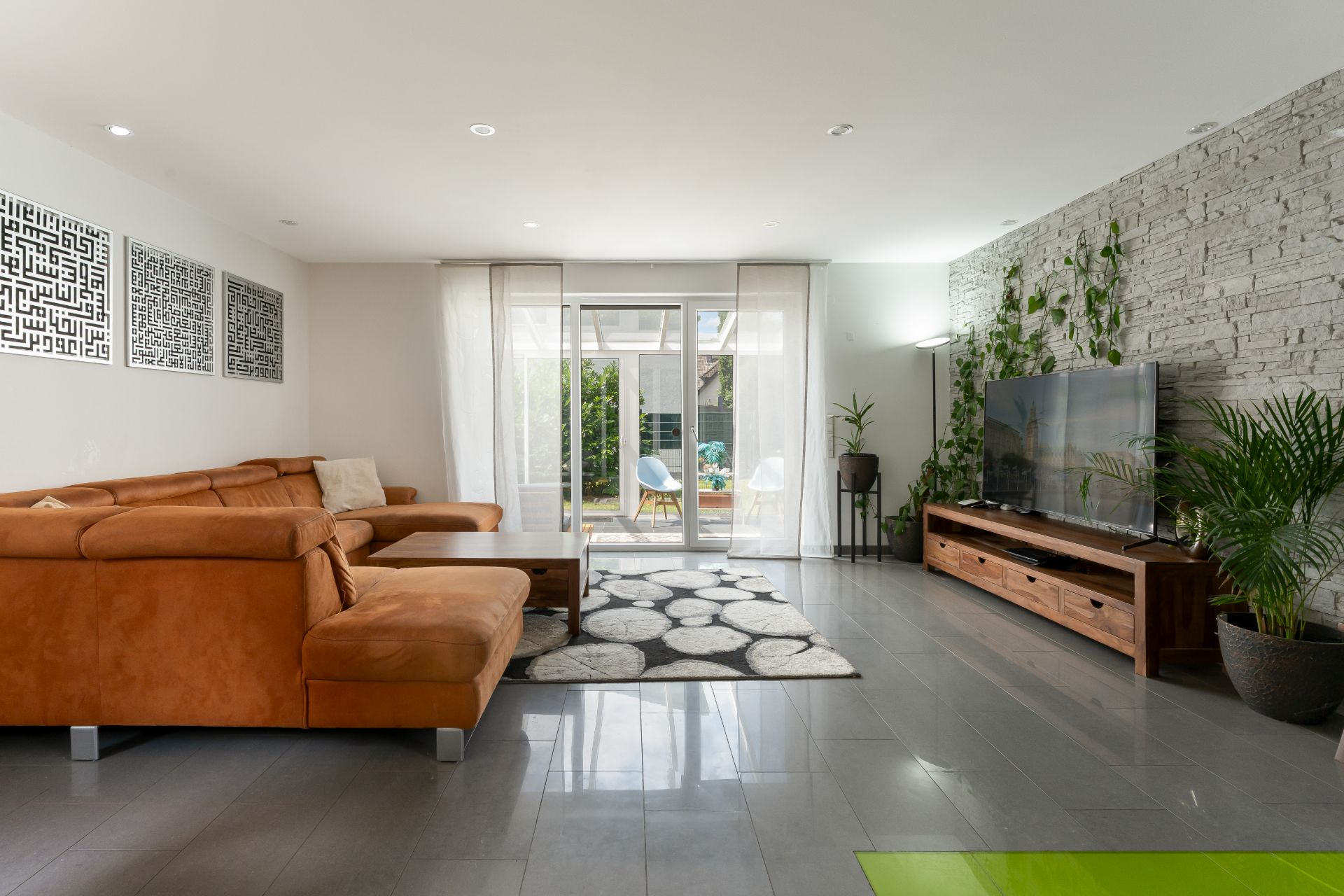 Modernes Wohnzimmer mit braunem Sofa, Teppich, Fernseher und Pflanzen, große Fenster zum Garten.