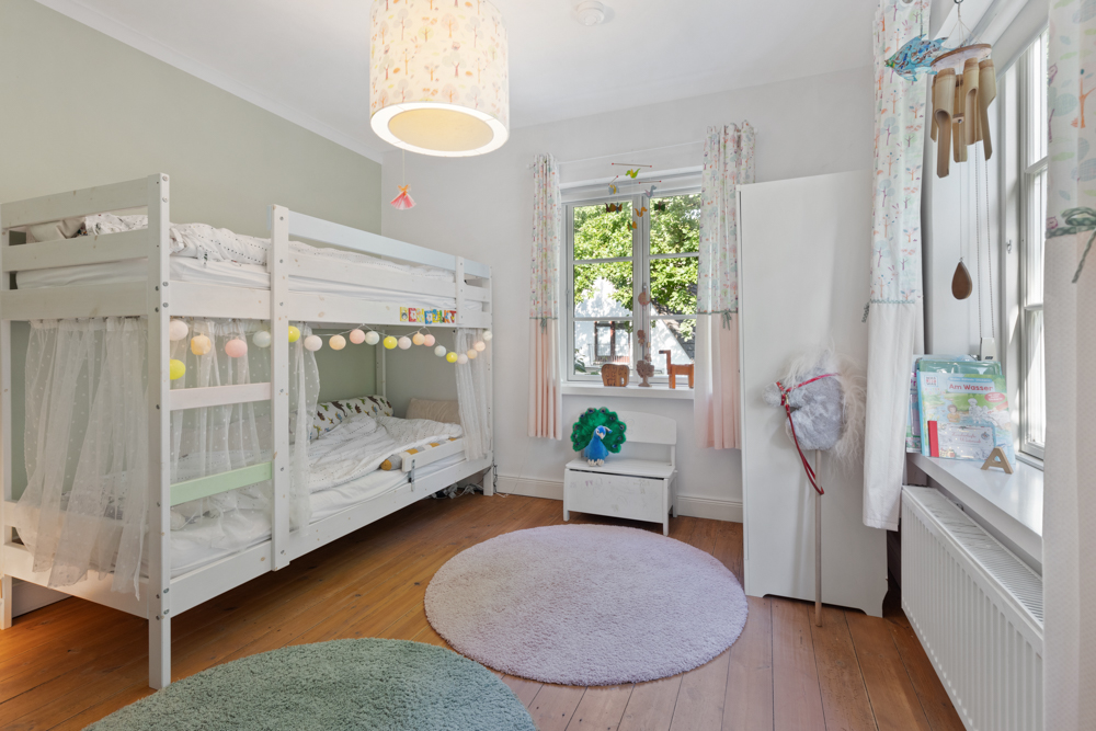 Helles Kinderzimmer mit Etagenbett, Teppichen, Schrank und Fenster mit Blick ins Grüne.