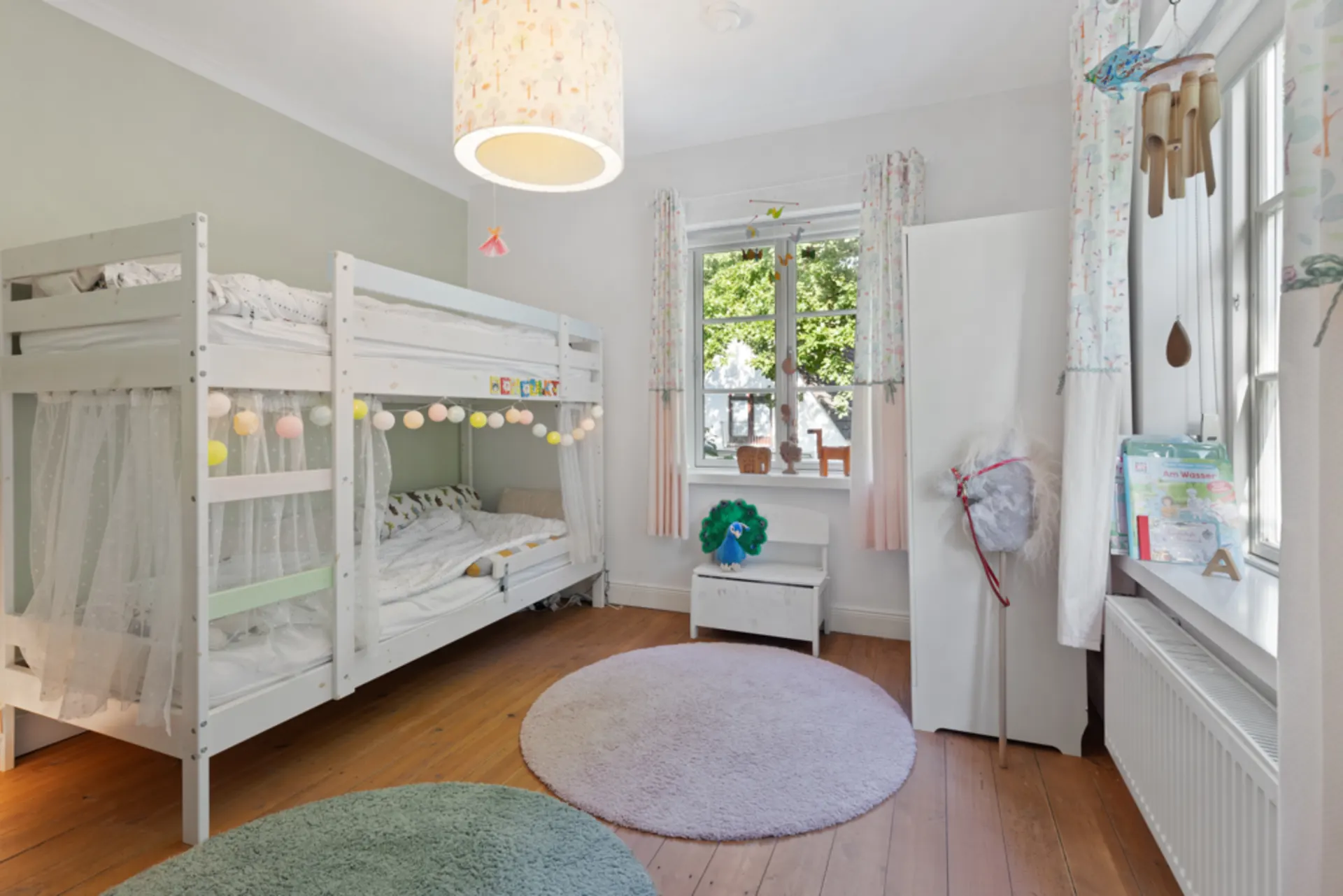 Helles Kinderzimmer mit Etagenbett, Teppichen, Schrank und Fenster mit Blick ins Grüne.