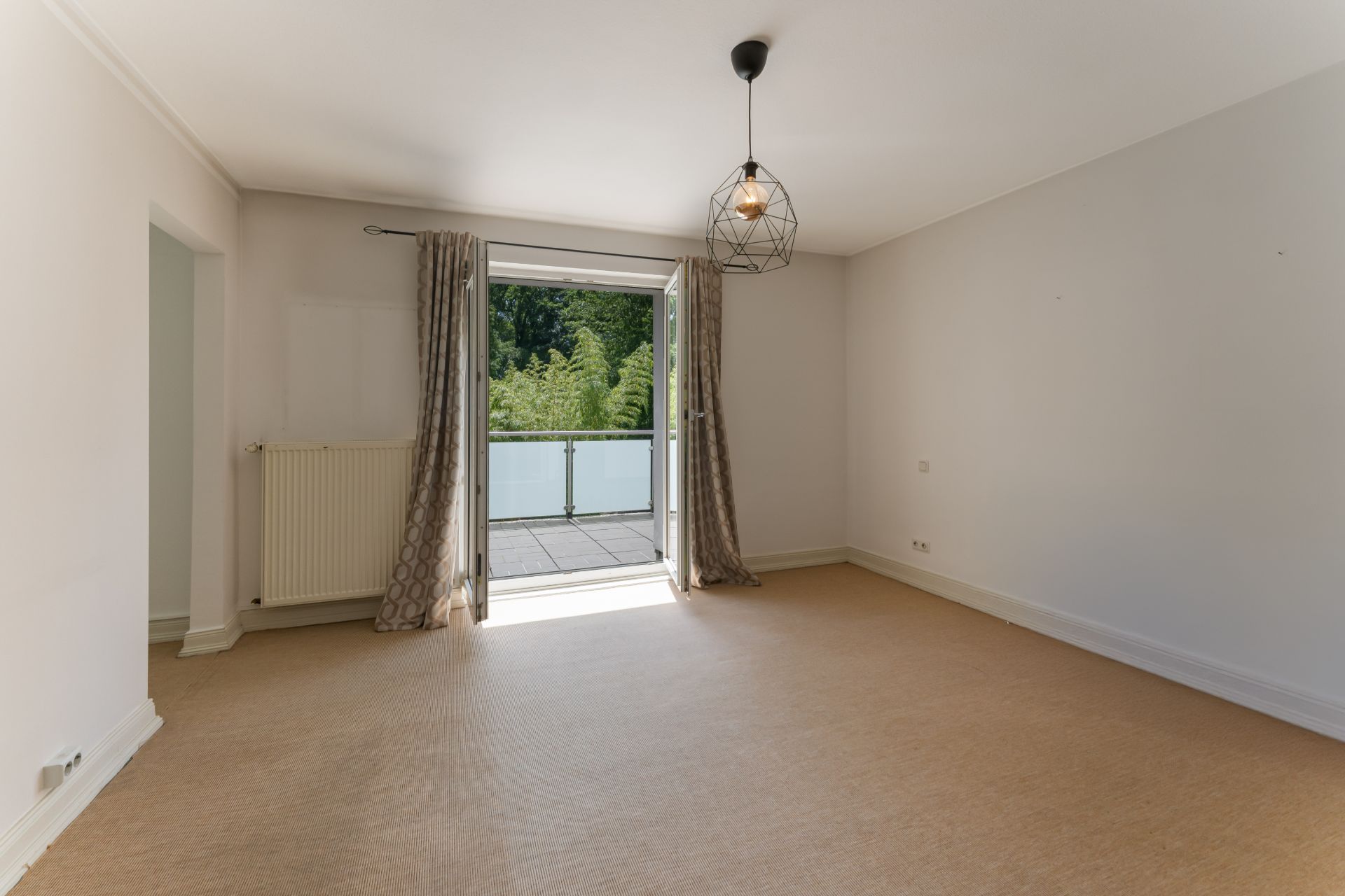 Helles, leeres Zimmer mit Teppichboden, Deckenlampe und Zugang zu einem Balkon mit Blick ins Grüne.