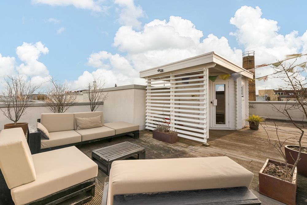 Dachterrasse mit hellen Sitzmöbeln, Holzdeck, kleinen Bäumen im Topf und weißem Gartenhaus.
