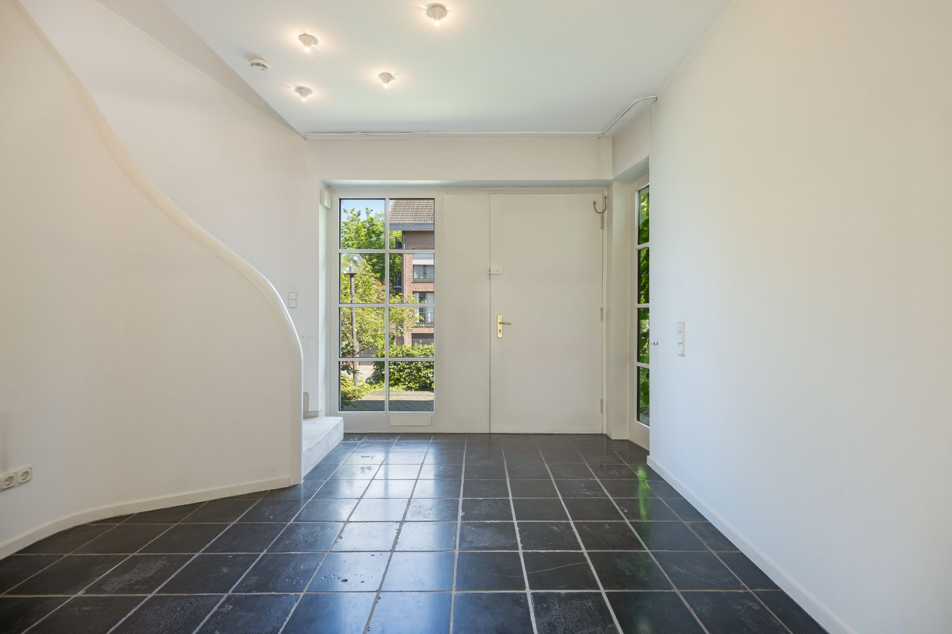 Heller Flur mit schwarzem Fliesenboden, weißer Treppe und einer Tür mit Glaseinsätzen.