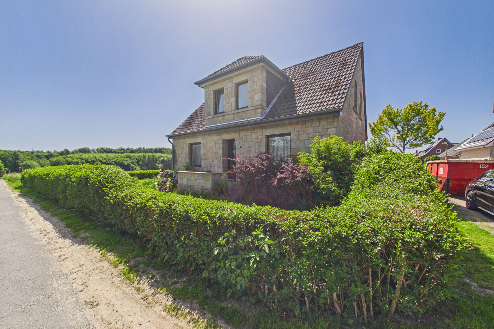 Steinhaus mit Satteldach, kleinen Büschen und Hecke entlang der Straße bei sonnigem Wetter.