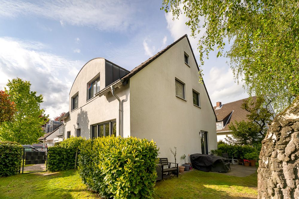 Zweistöckiges Wohnhaus mit Garten, Terrasse und Gartenmöbeln, umgeben von Büschen und Bäumen.