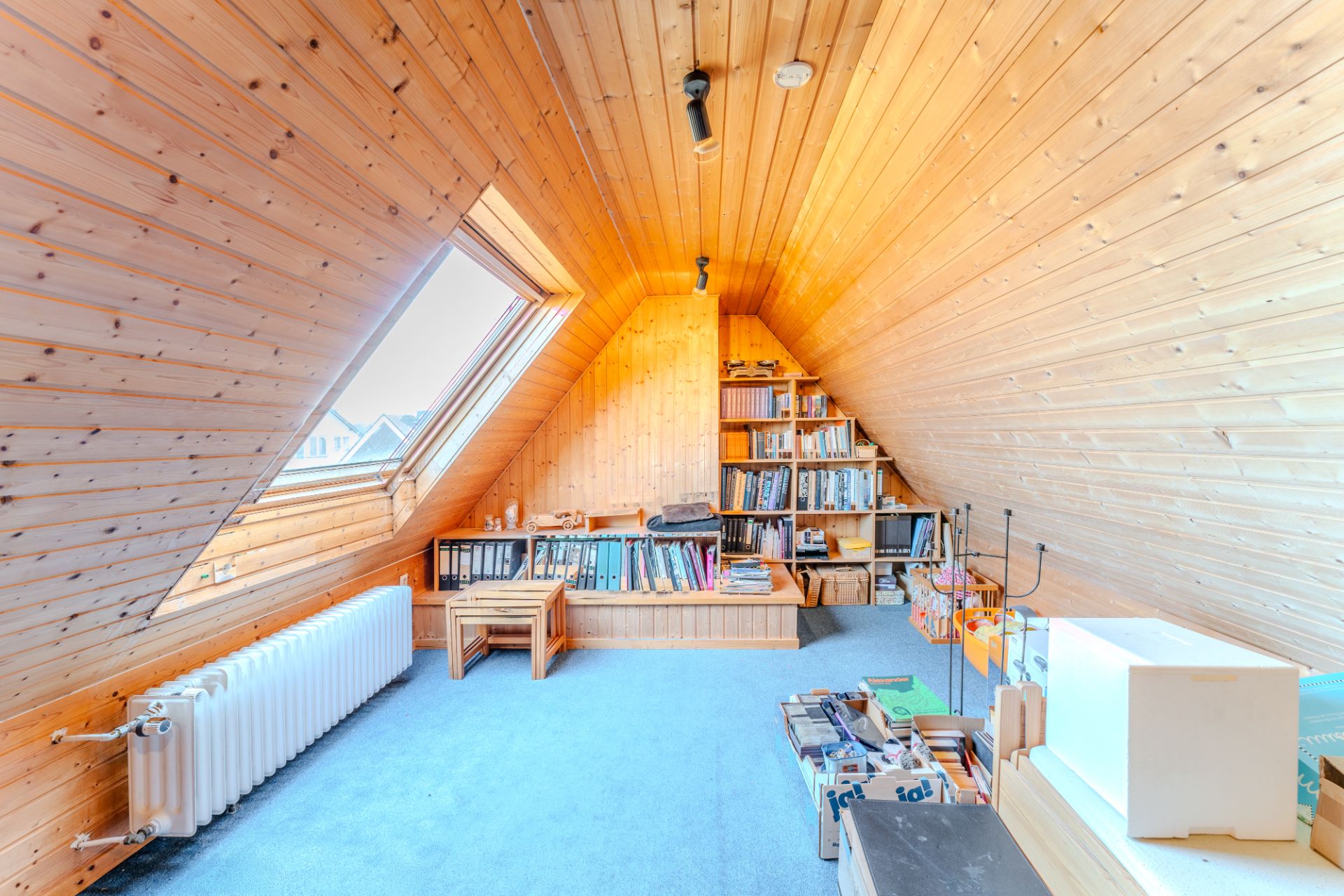 Heller Dachbodenraum mit Holzverkleidung, Bücherregalen, Schreibtisch und schrägem Fenster.