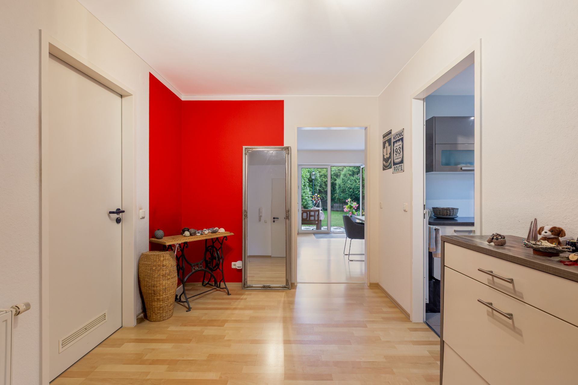 Heller Flur mit roter Wand, Spiegel, Tisch, Zugang zu Küche und Blick ins Wohnzimmer mit Gartenausgang.