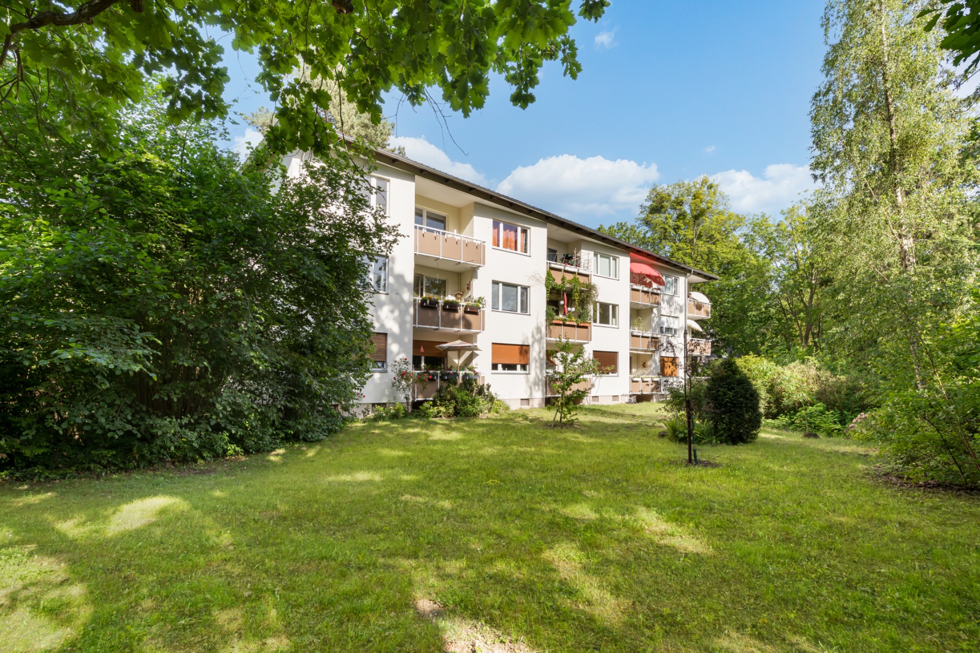 Mehrfamilienhaus mit Balkonen und großem Garten, umgeben von Bäumen und Rasen.