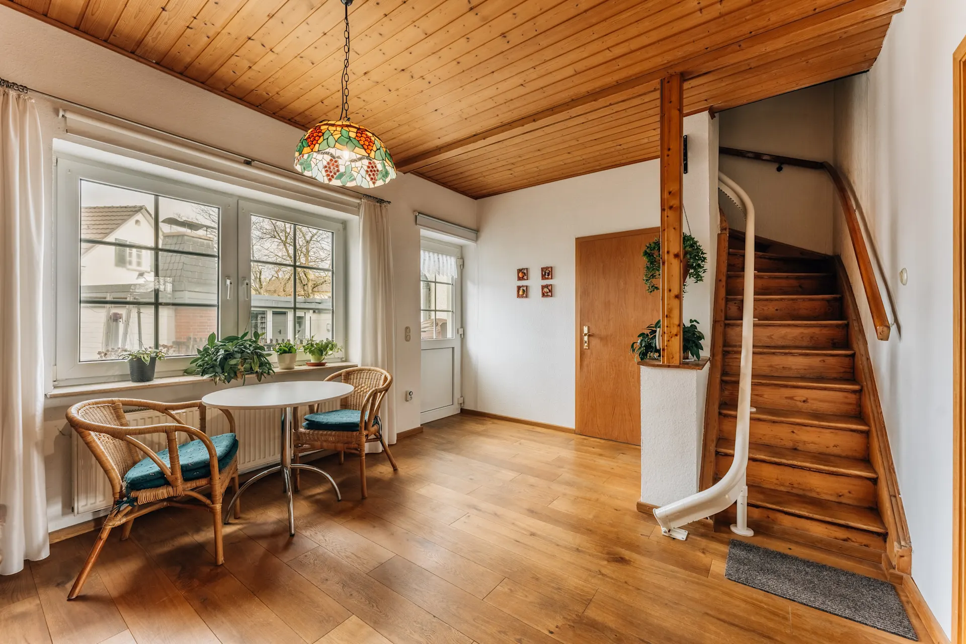 Heller Raum mit Holzdecke, Treppe, Tisch mit zwei Stühlen und Pflanzen am Fenster.