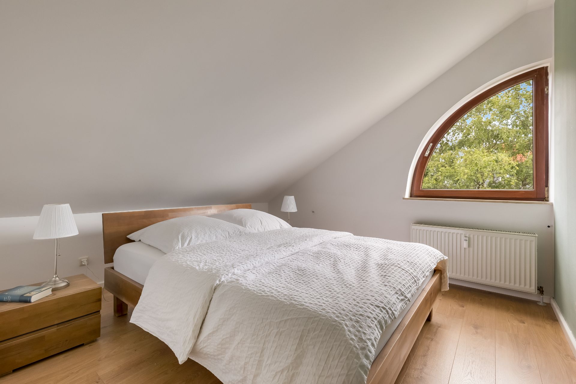 Schlafzimmer mit schräger Decke, Holzbett, Nachttisch und großem halbrunden Fenster.
