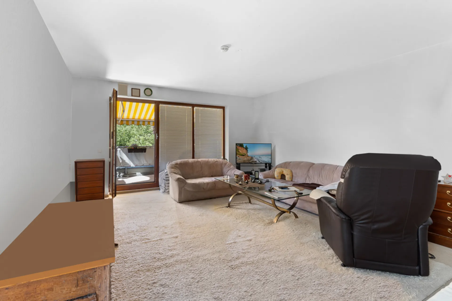 Wohnzimmer mit Sofa, Sessel, Fernseher und Zugang zu einem Balkon mit Markise.
