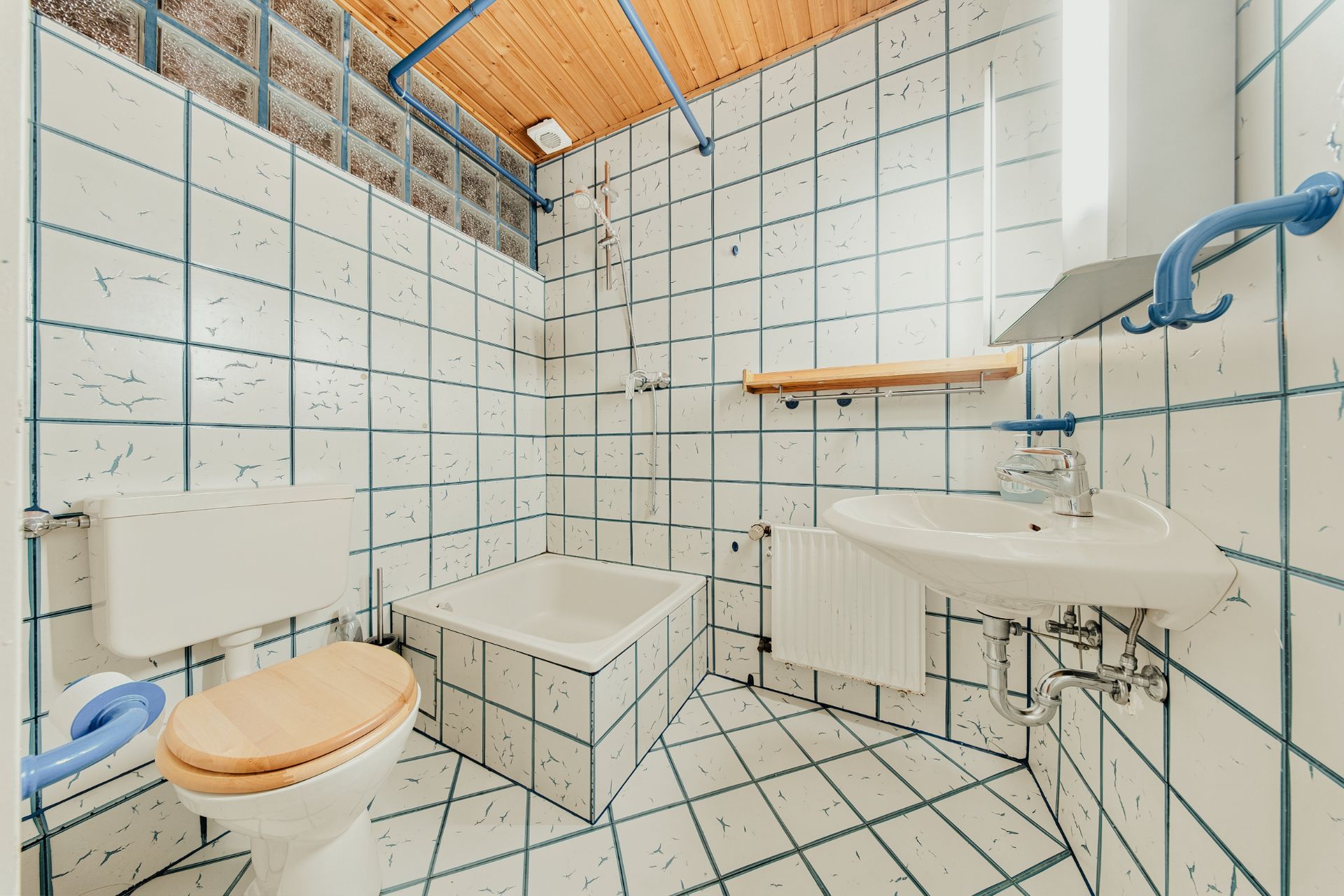 Badezimmer mit Toilette, kleinem Waschbecken, Badewanne und weißen Fliesen mit blauen Linien.