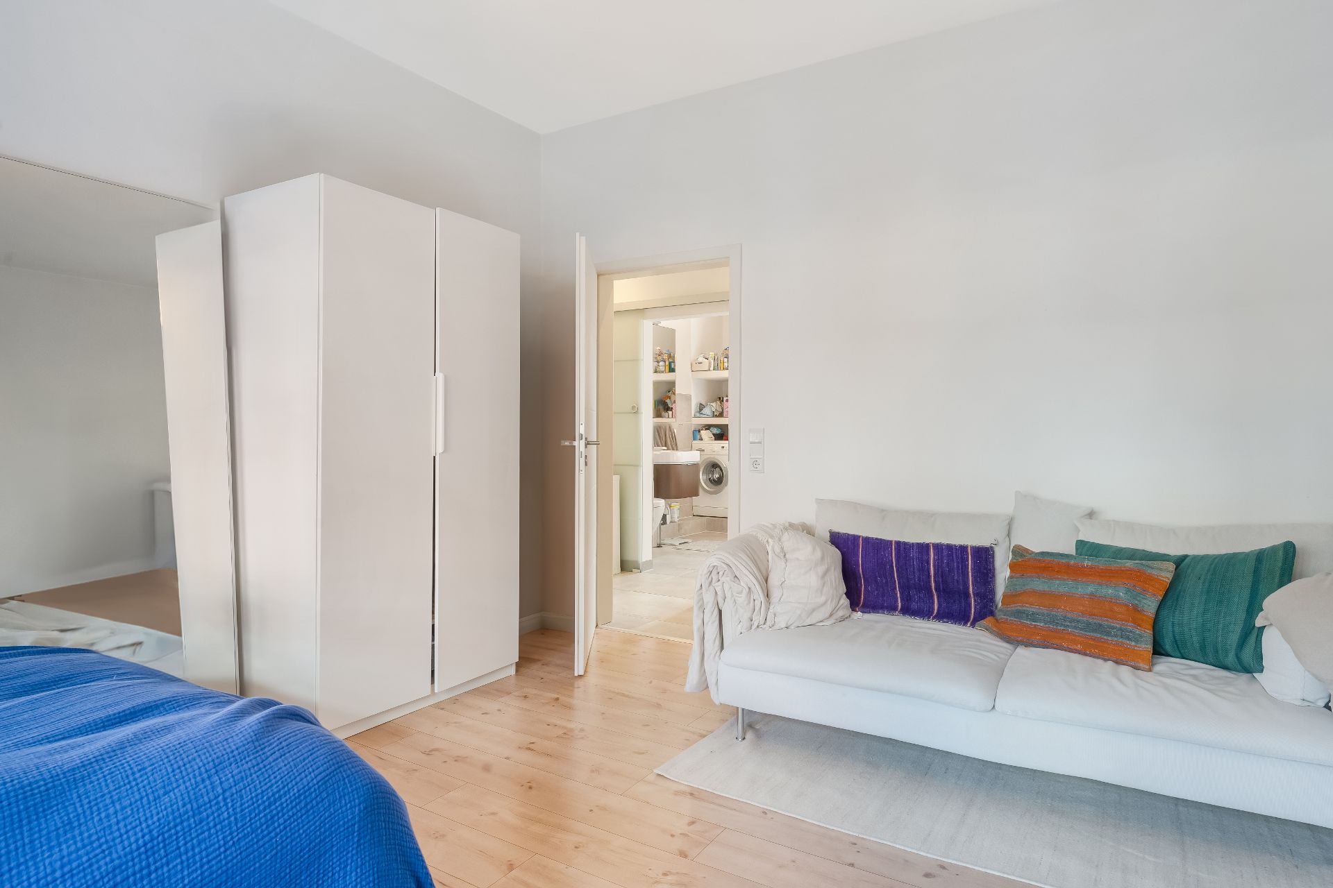 Helles Zimmer mit weißem Kleiderschrank, Sofa mit bunten Kissen und offenem Türdurchgang.