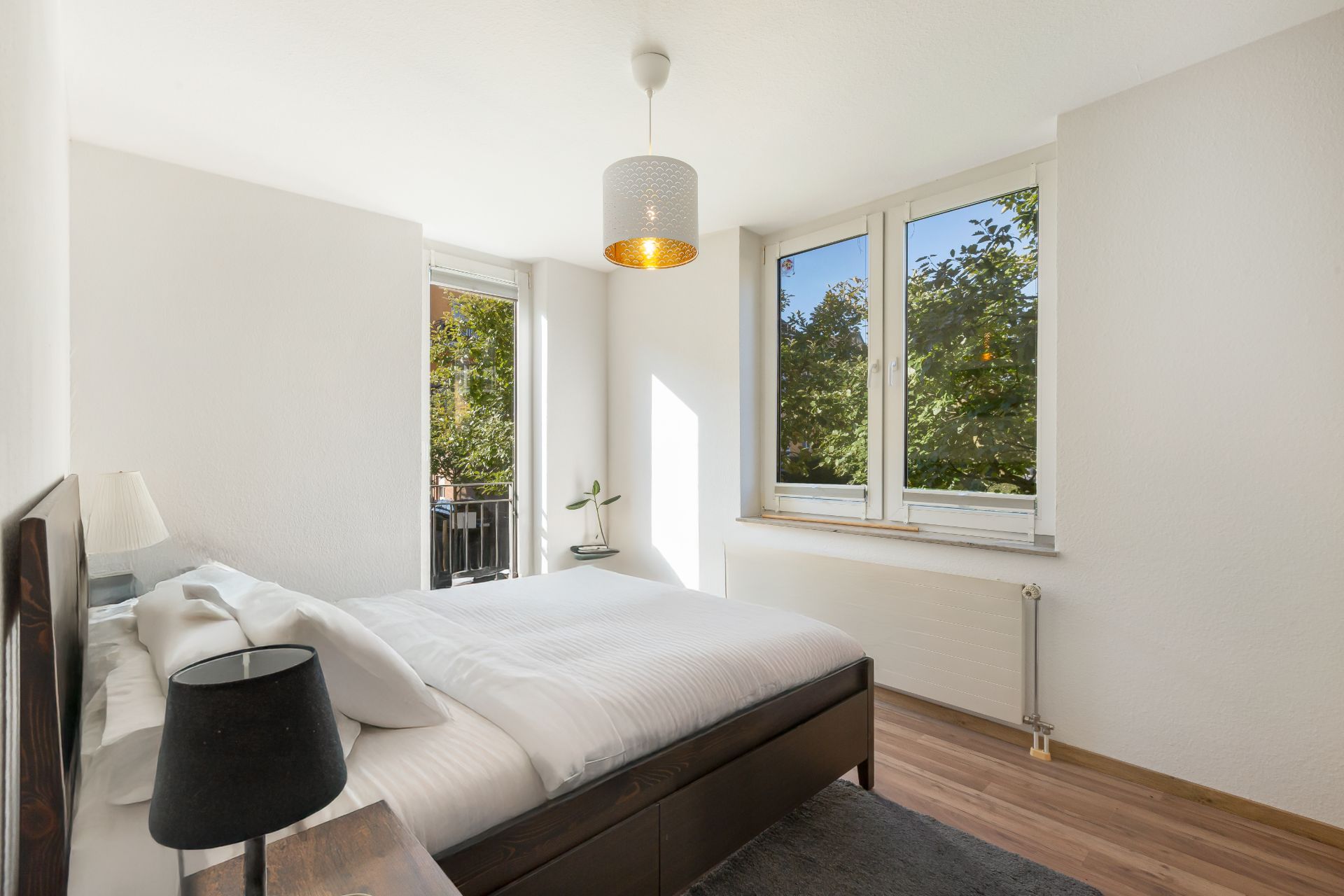 Helles Schlafzimmer mit Doppelbett, Nachttischen, Lampe und großen Fenstern mit Blick auf grüne Bäume.