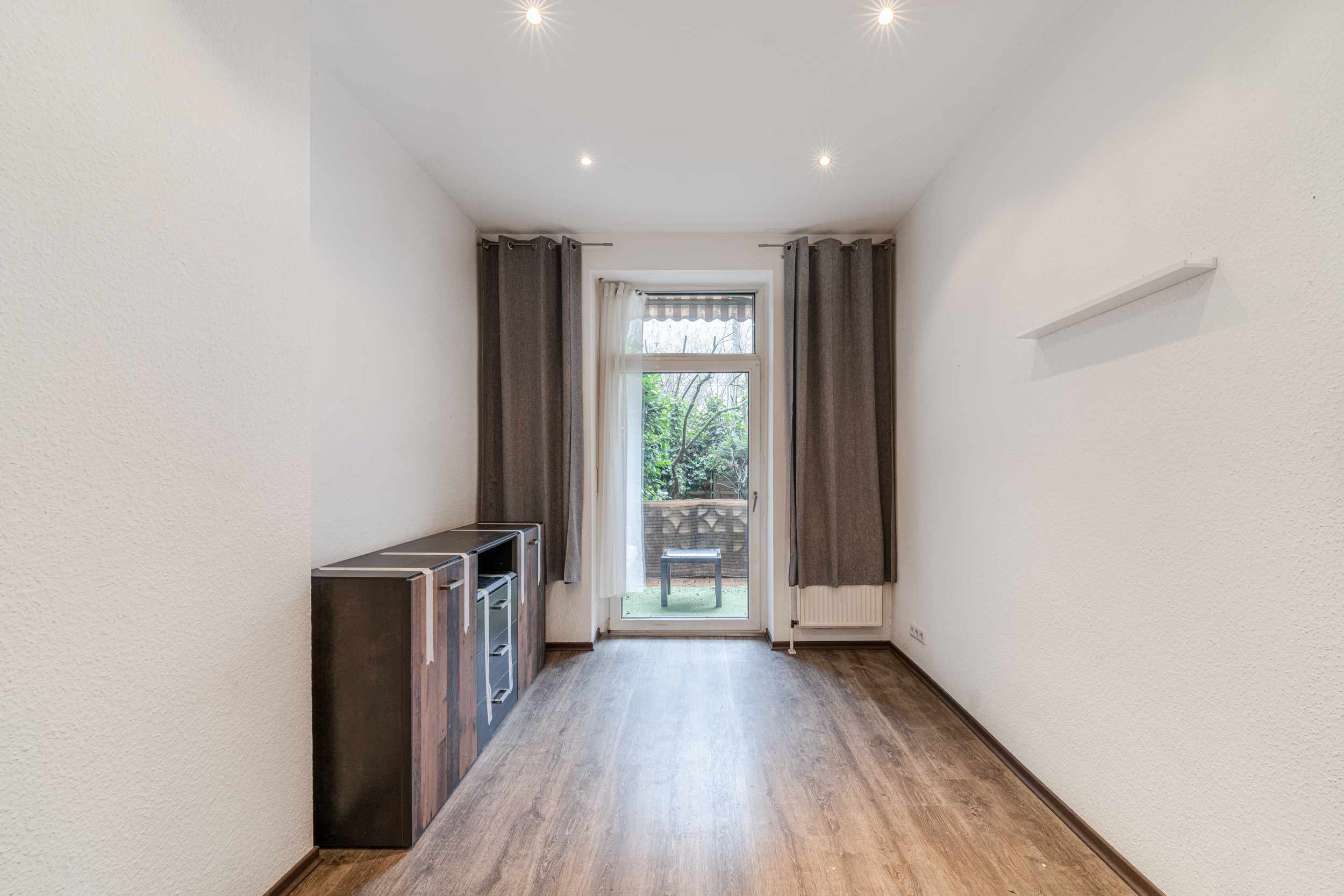 Leerer Raum mit Holzboden, Fenster mit Vorhängen und kleinem Regal, Blick auf Terrasse.