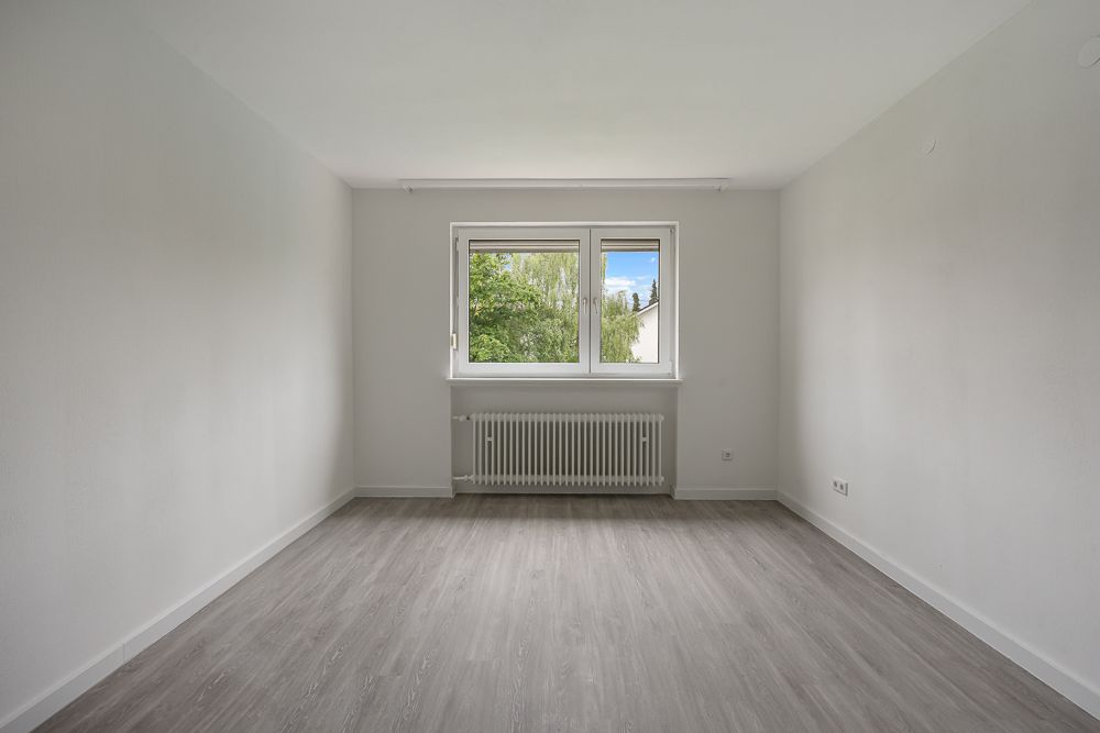 Leerer Raum mit grauem Boden, weiß gestrichenen Wänden und großem Fenster mit Blick ins Grüne.