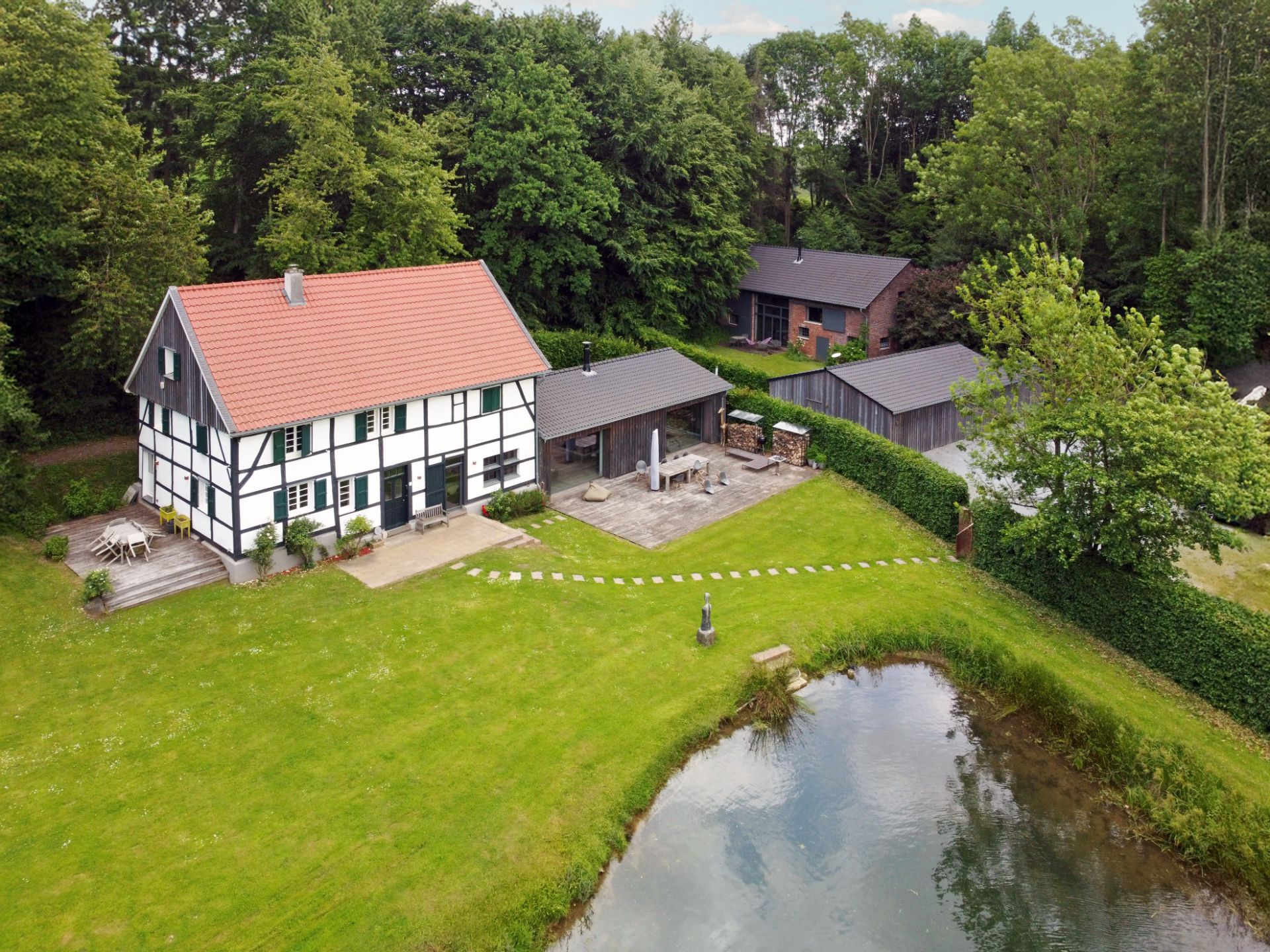 Fachwerkhaus mit Garten, Terrasse, Teich und Nebengebäuden, umgeben von Bäumen.