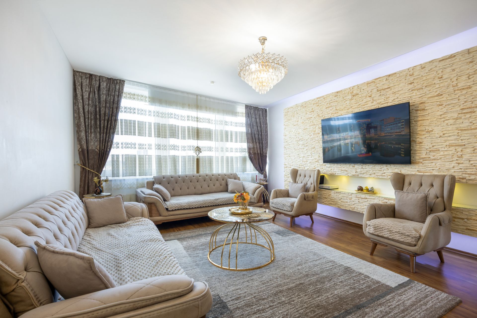 Modernes Wohnzimmer mit beigen Sofas, zwei Sesseln, Couchtisch, Fernseher und großem Fenster.