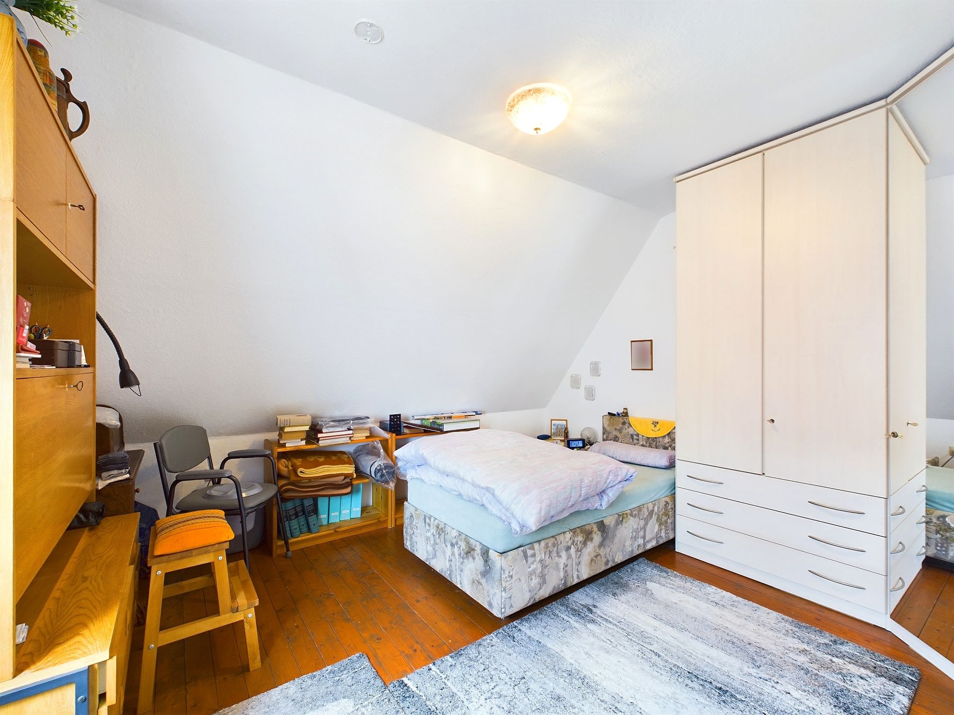 Schlafzimmer mit Bett, großem Kleiderschrank, Regal, Stühlen und Nachttisch unter Dachschräge.