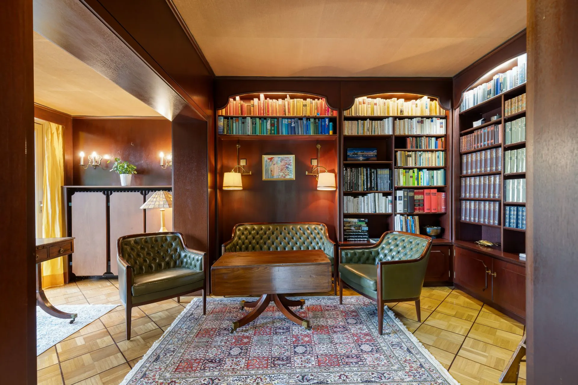 Wohnzimmer mit Bücherregalen, drei grünen Sesseln und einem Holztisch auf einem Teppich.
