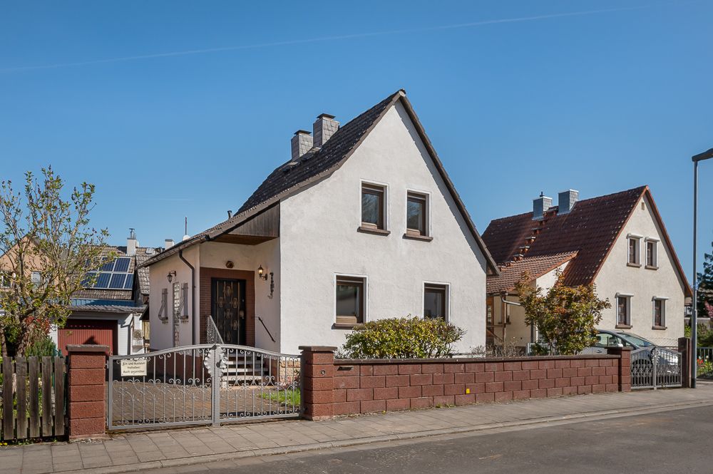 Weißes Einfamilienhaus mit dunklem Dach und Vorgarten hinter einem Zaun an einer Straße.