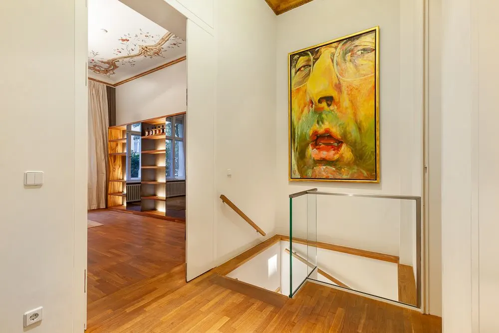 Flur mit Holzfußboden, Treppenabgang, großem Porträtgemälde und Blick in ein Zimmer mit Regal.