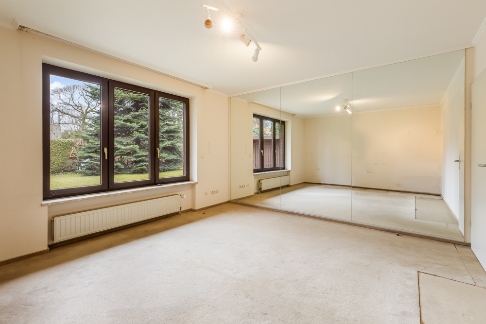 Leerer Raum mit Teppichboden, großem Fenster und Wandspiegel, Blick auf Garten.