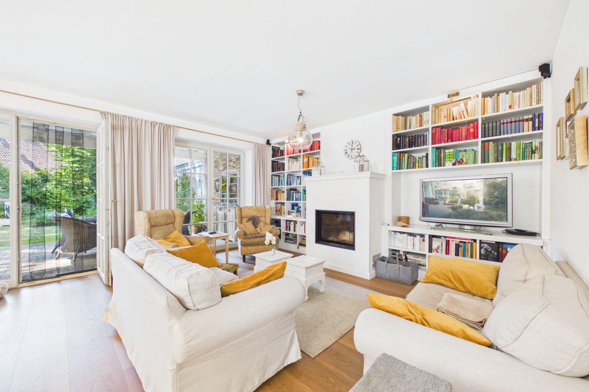 Helles Wohnzimmer mit Sofas, Kamin, Fernseher, Bücherregalen und Zugang zum Garten.