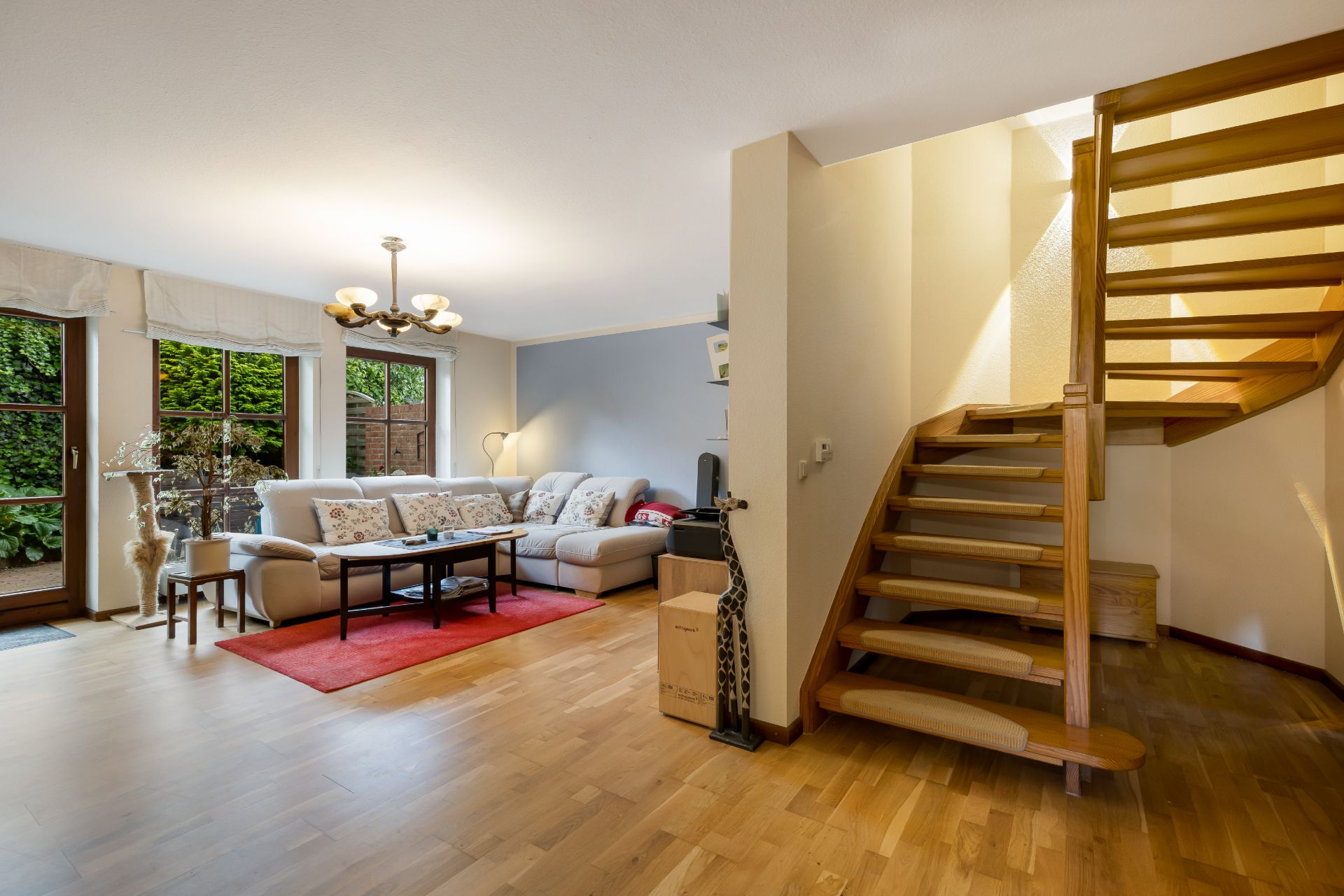 Helles Wohnzimmer mit Ecksofa, rotem Teppich, Holzboden und offener Holztreppe.
