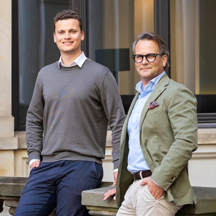 Zwei Männer stehen vor einem Fenster eines Wohngebäudes und lächeln in die Kamera.