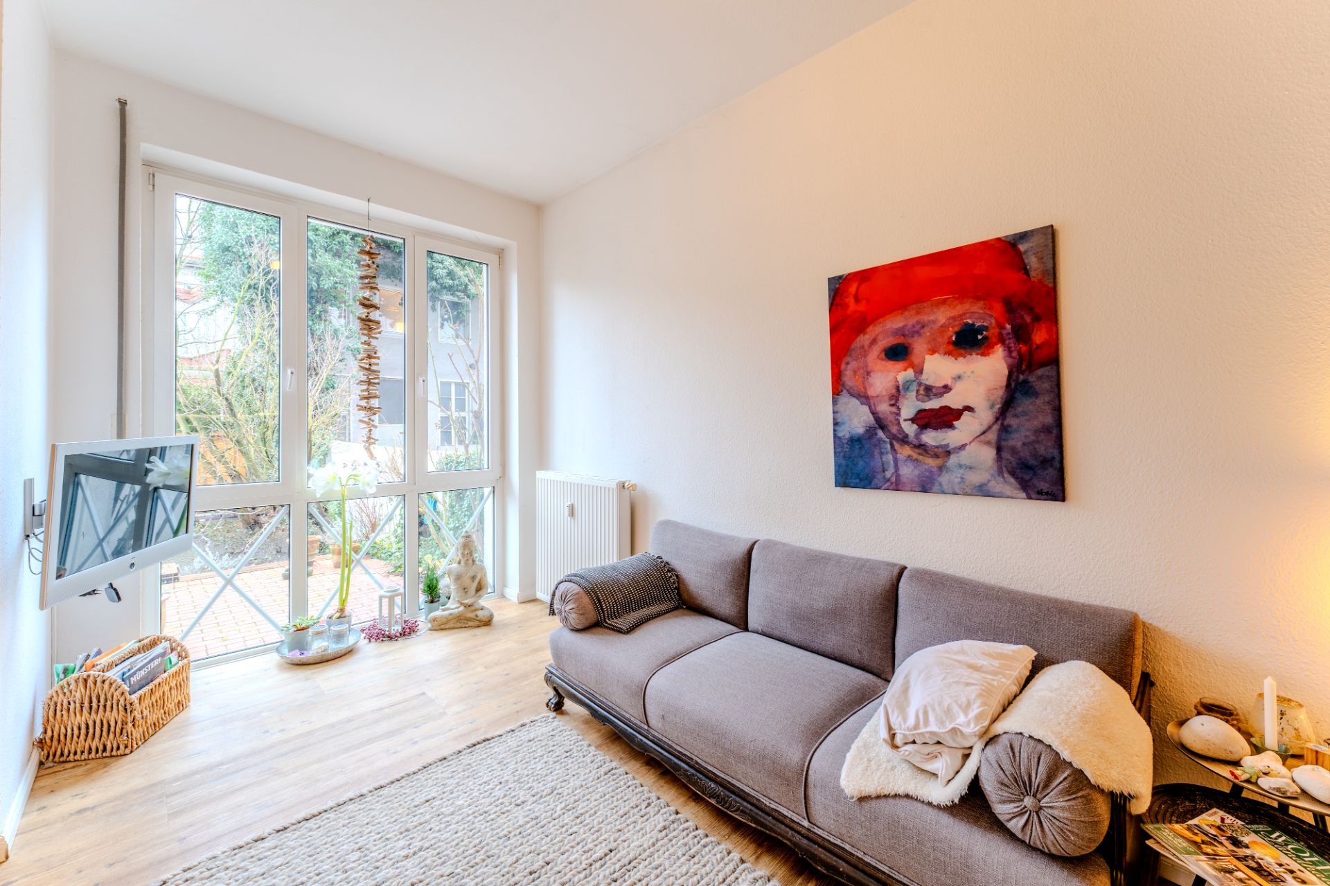 Helles Wohnzimmer mit grauem Sofa, großem Fenster und buntem Porträtbild an der Wand.