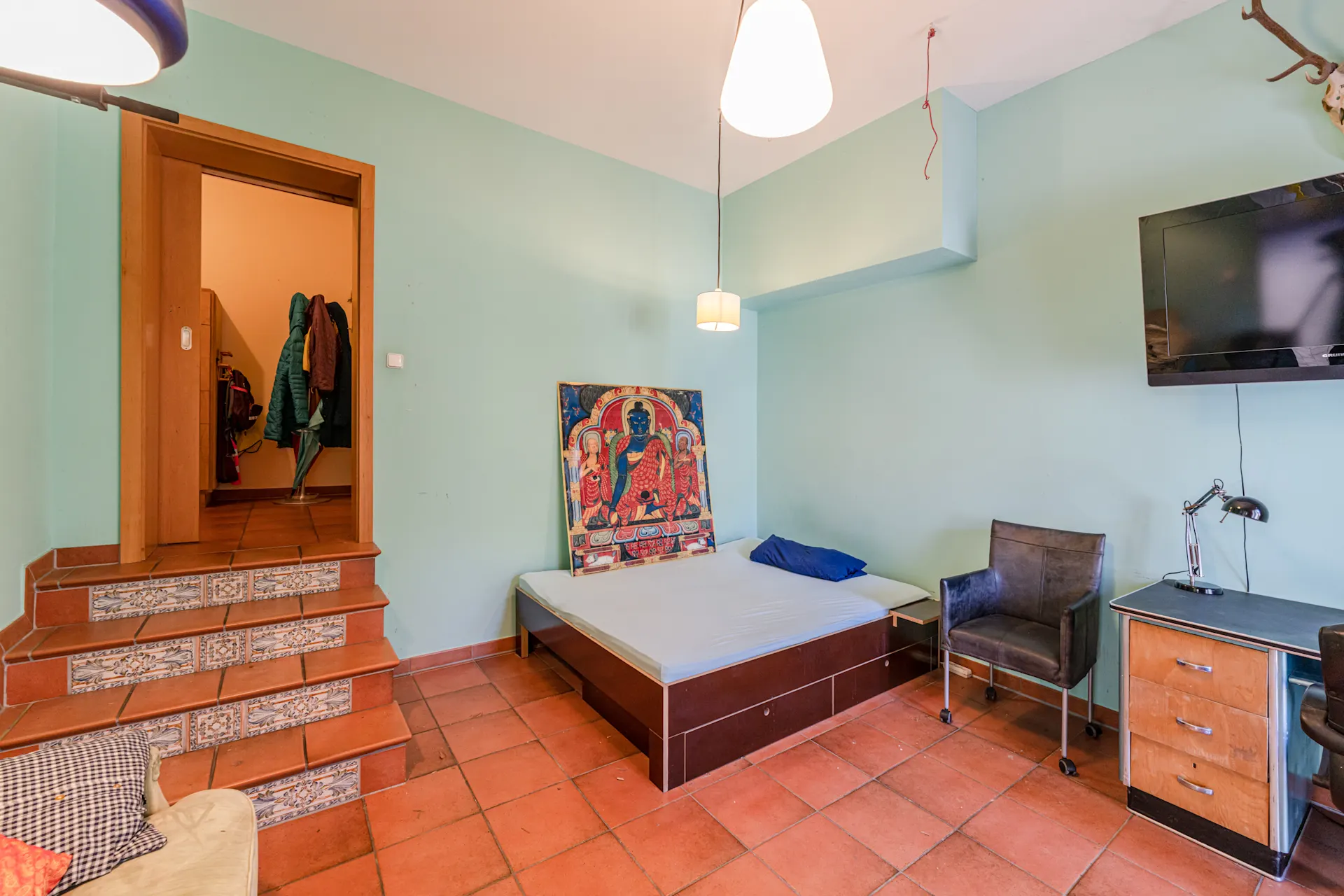 Zimmer mit Bett, Schreibtisch, Stuhl und Wandbild, Treppe führt zu einem Flur mit Garderobe.