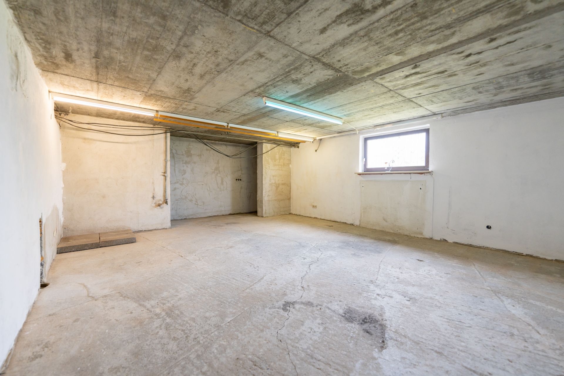Leerer Kellerraum mit Betonboden, weiß gestrichenen Wänden und einem kleinen Fenster.