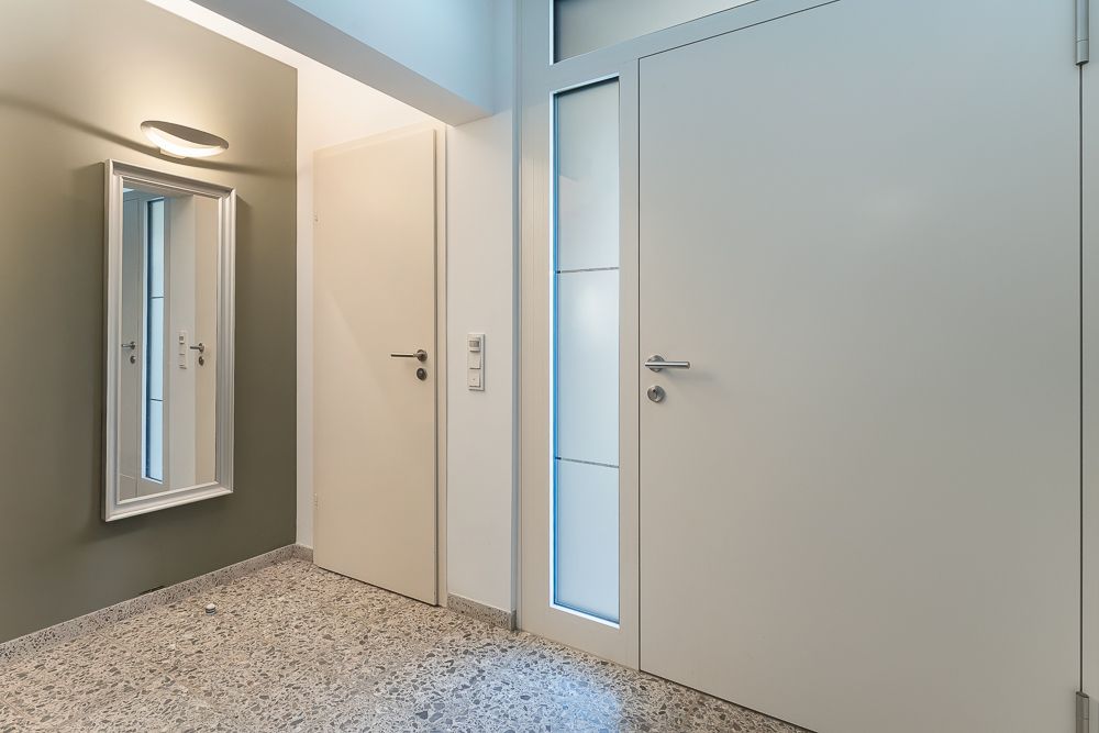 Heller Flur mit zwei weißen Türen, einem Wandspiegel und grauem Boden.