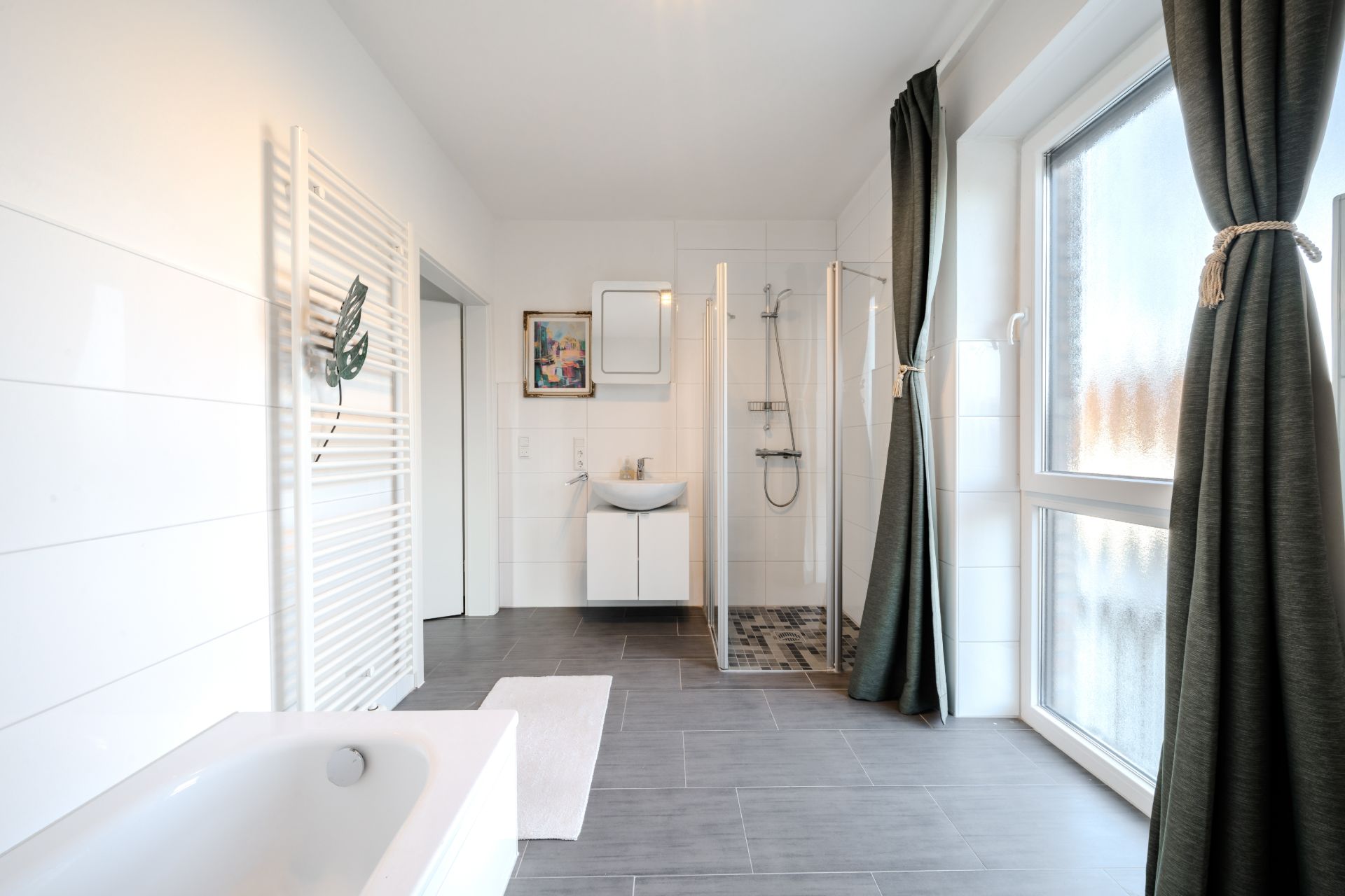 Helles Badezimmer mit Badewanne, Dusche, Waschbecken und großen Fenstern mit Vorhängen.