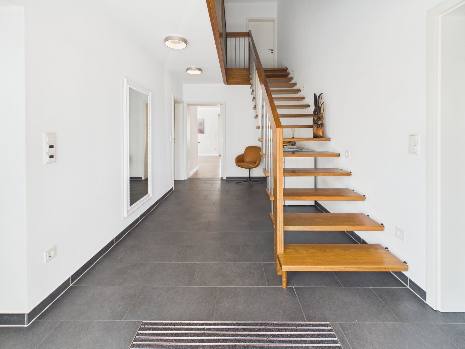 Heller Flur mit Holztreppe, großem Spiegel und grauen Fliesen.