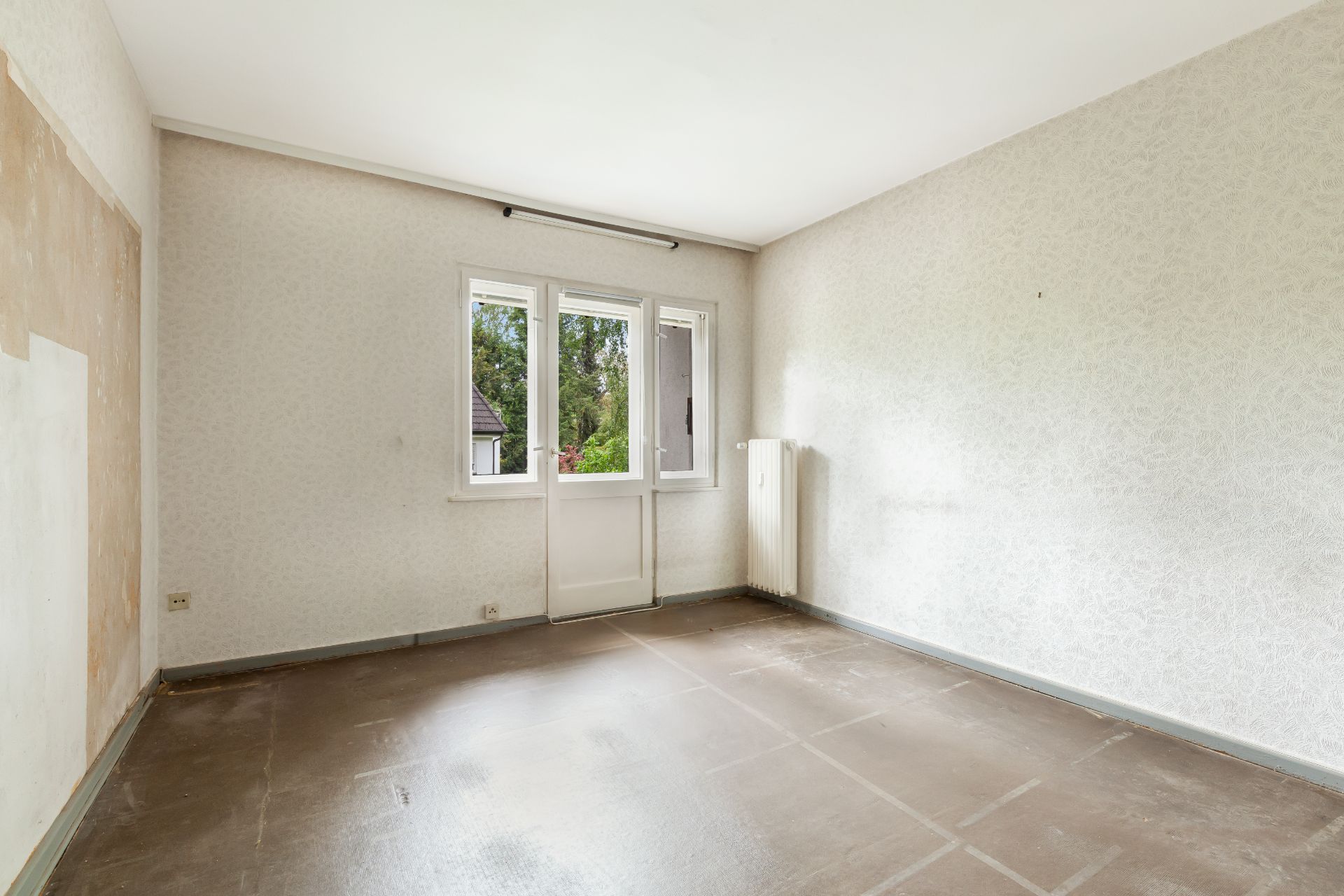 Leeres Zimmer mit Fenster, Tür zum Balkon und Heizkörper, helle Wände und brauner Boden.