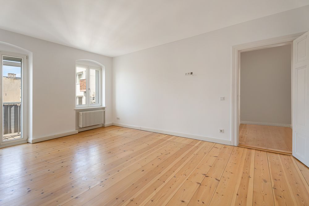 Leeres Zimmer mit Holzfußboden, weißen Wänden und zwei Fenstern, eines mit Zugang zum Balkon.