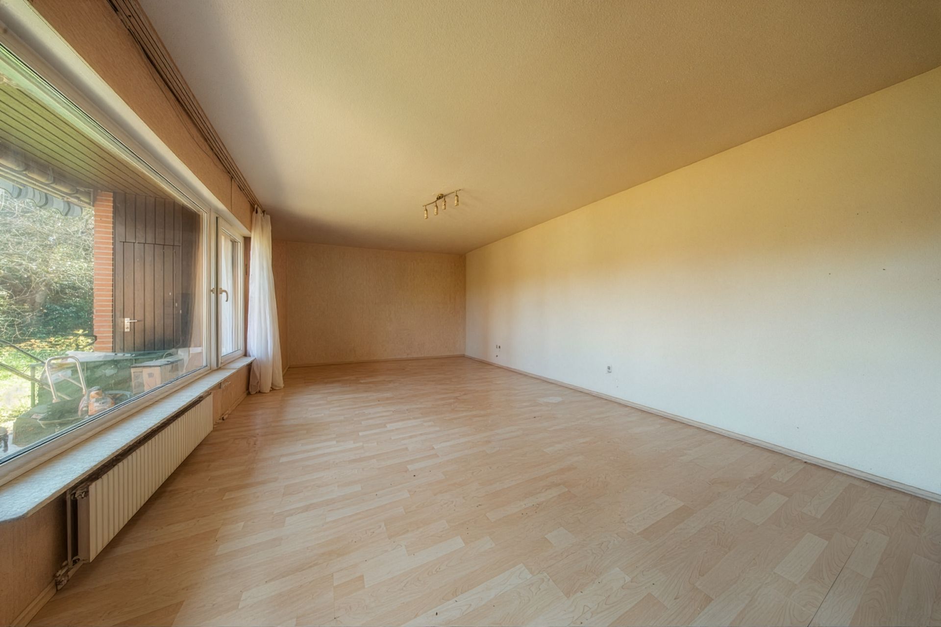 Leeres Wohnzimmer mit großem Fenster, Parkettboden und Blick in den Garten.