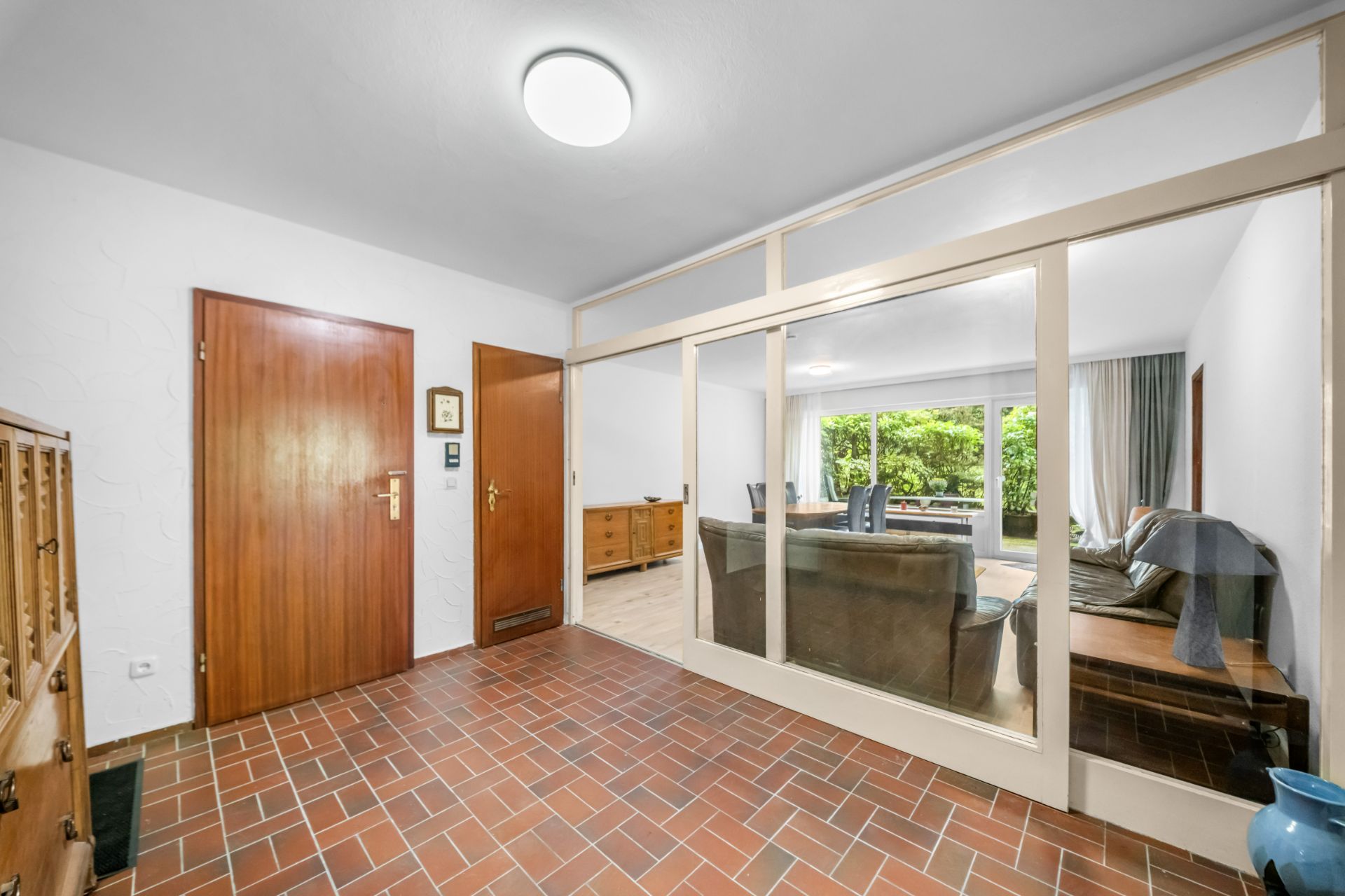 Heller Flur mit Fliesenboden, Holztüren und Glaswand zu einem Wohnzimmer mit Sofa und großem Fenster.