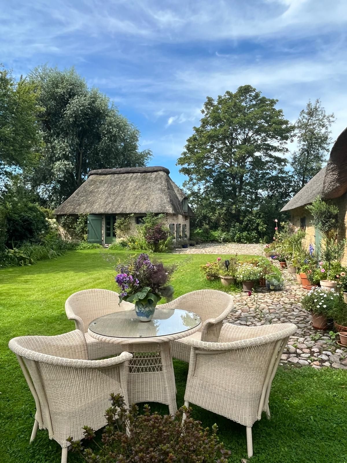 Garten mit rundem Glastisch, vier Korbstühlen und reetgedecktem Haus im Hintergrund.