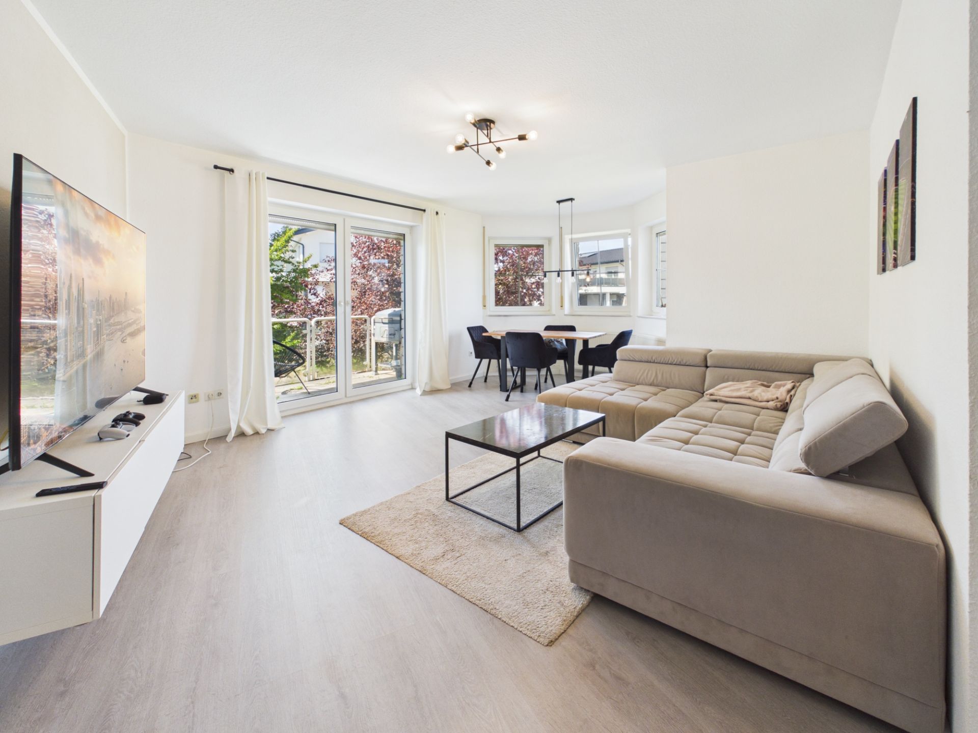 Helles Wohnzimmer mit Ecksofa, Couchtisch, großem Fernseher und Essbereich am Fenster.