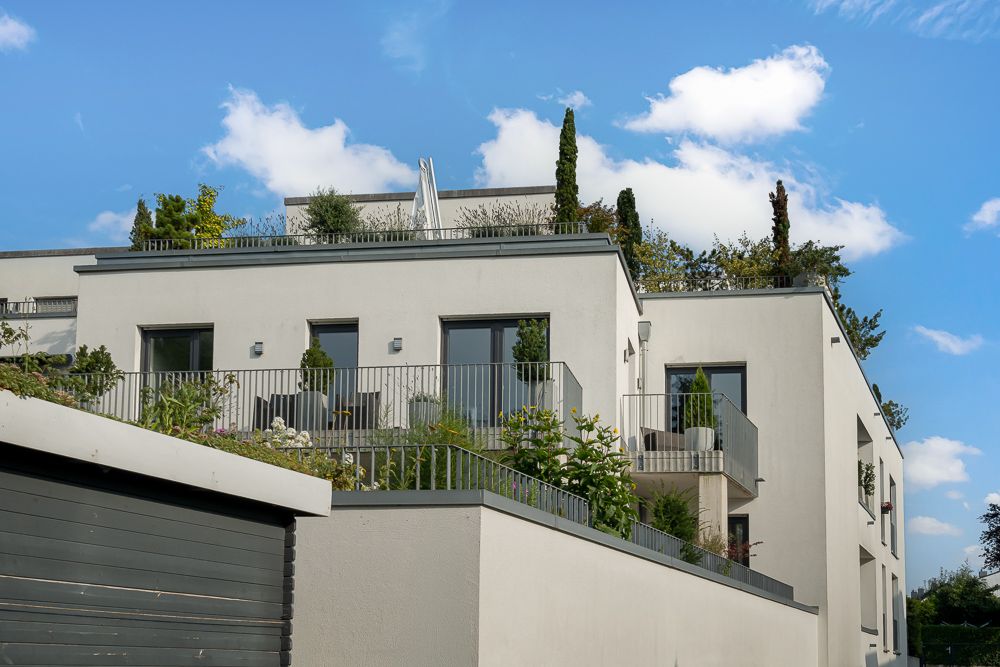 Modernes weißes Wohngebäude mit Balkonen, Pflanzen und Flachdach bei blauem Himmel.