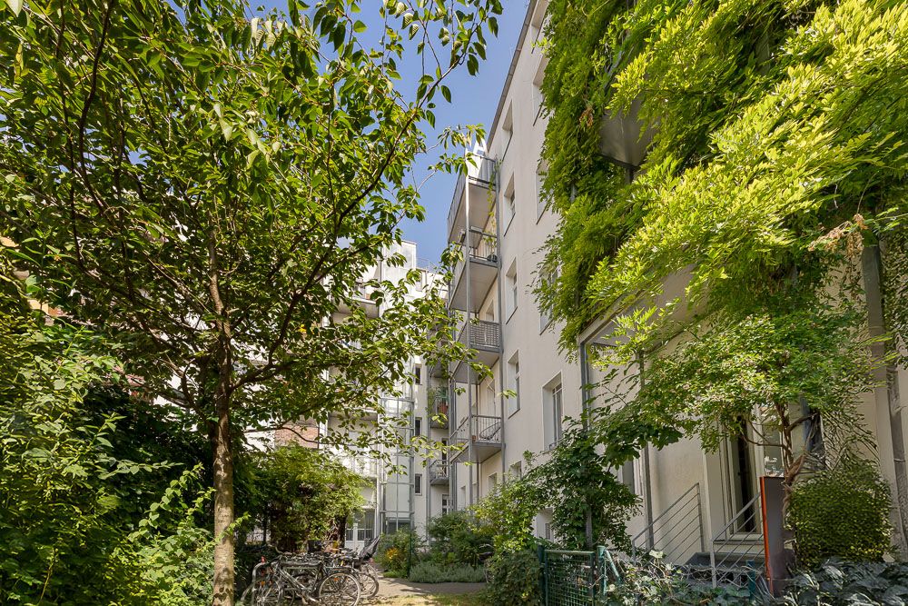 Mehrstöckiges Wohnhaus mit Balkonen und begrüntem Hof, Bäume und Fahrräder im Vordergrund.