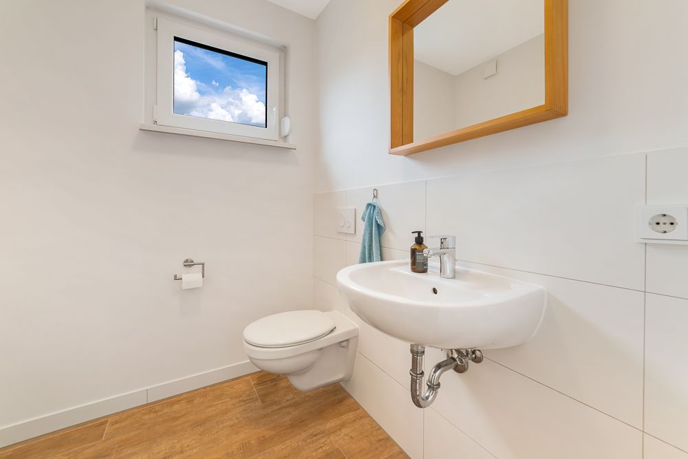 Helles Badezimmer mit Wandtoilette, Waschbecken, Spiegel und Fenster.