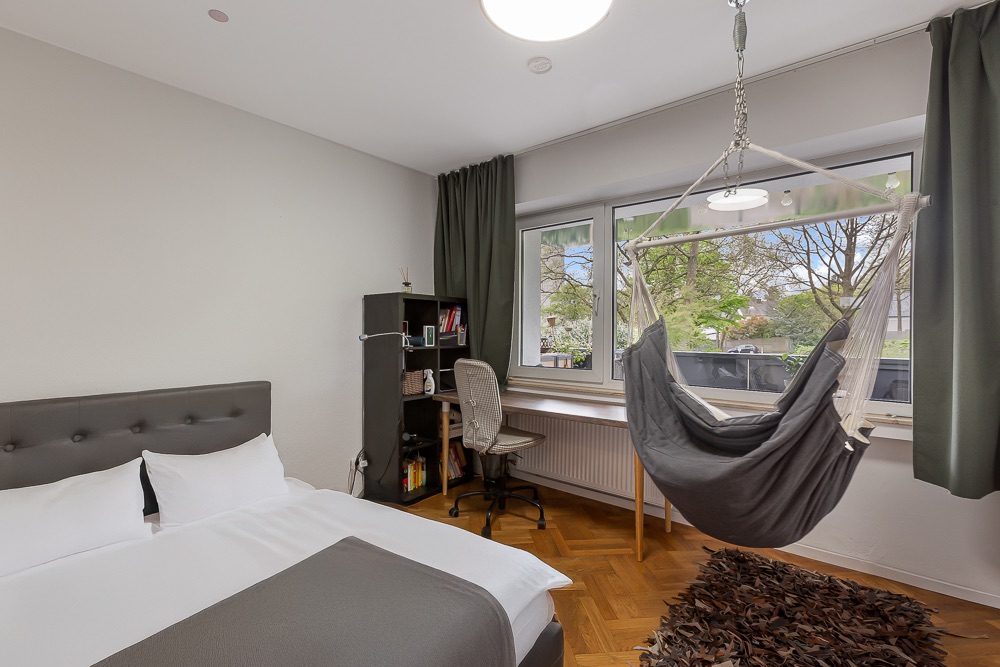 Schlafzimmer mit Bett, Schreibtisch, Bücherregal und Hängesessel vor einem Fenster.