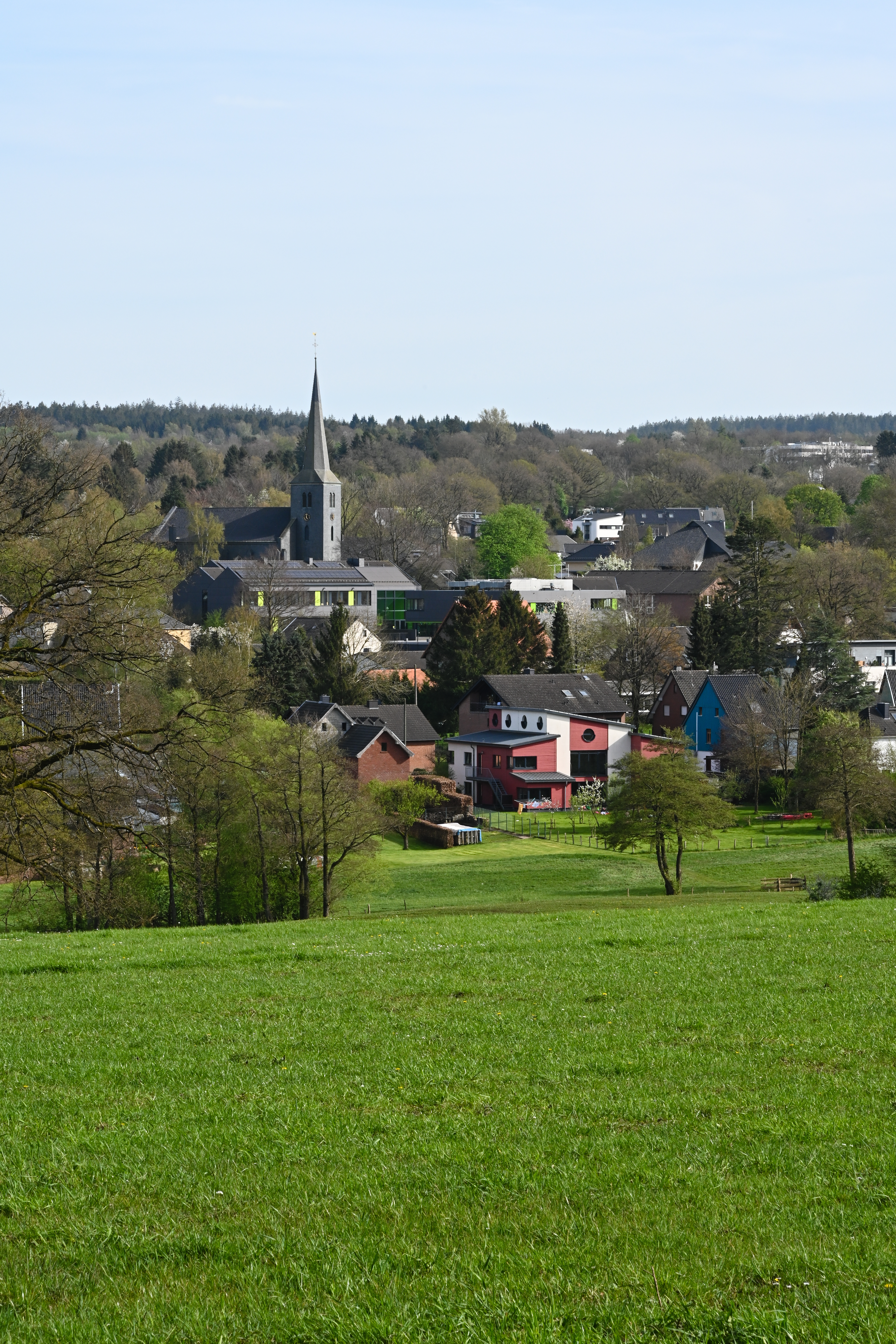 Luftaufnahme eines Wohngebiets mit Häusern, Feldern und Windrädern im Hintergrund.